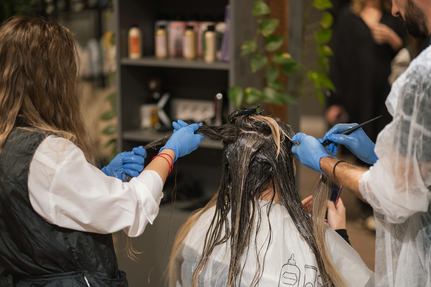 Two hairstylists in blue gloves dye a client's wet hair.